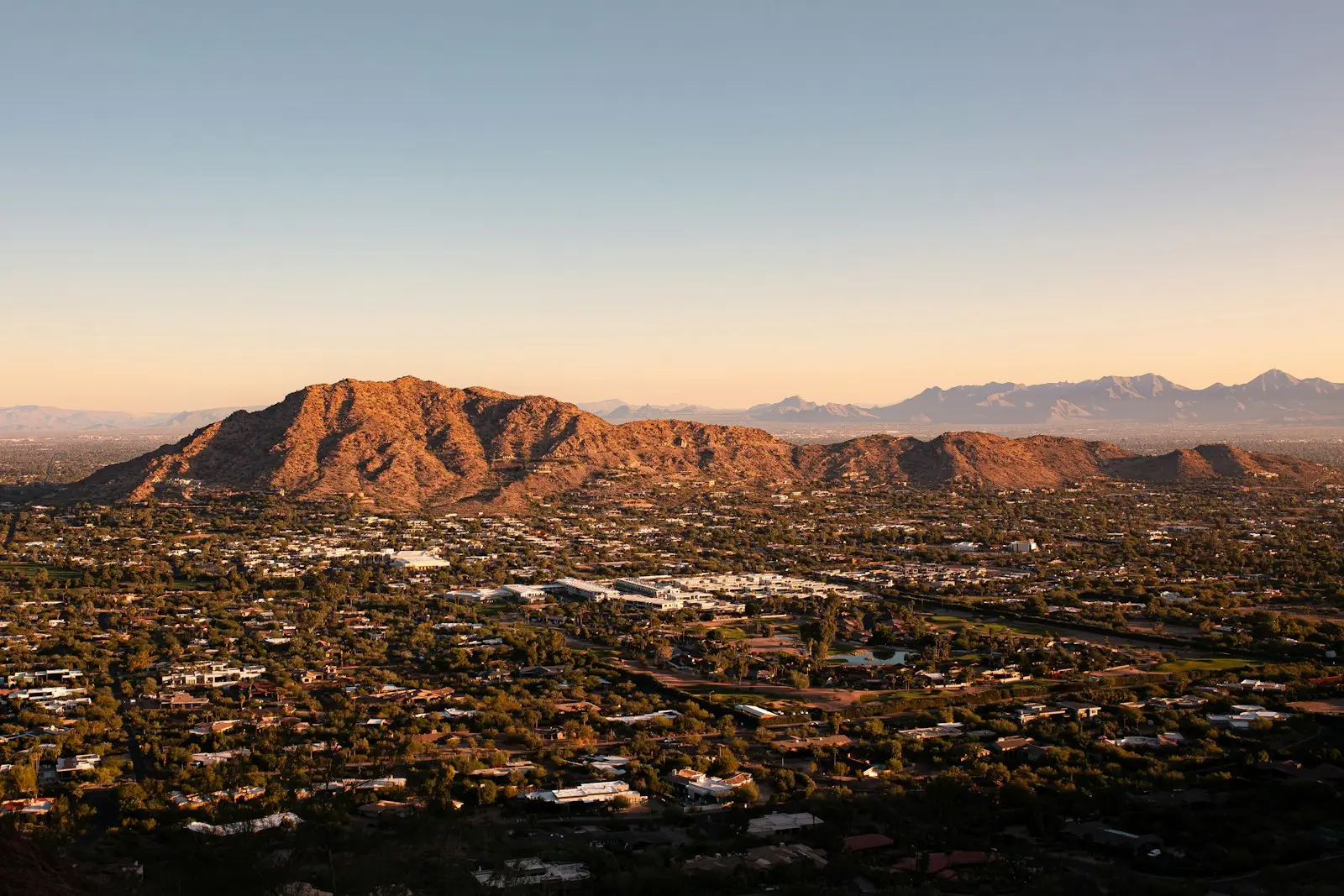 A rocky desert mountain rising over a valley of low homes and trees at golden hour
